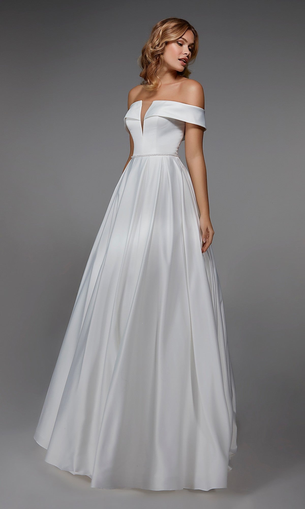 A woman wears the Long White Dress: Alyce 7023, featuring an off-the-shoulder fitted bodice and a floor-length skirt, standing against a plain gray background.